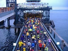 A large group of cyclists, wearing colorful attire, rides onto a ferry over a boarding ramp above the water.