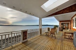 A wooden deck with chairs and a table overlooks a calm body of water. The deck is covered with a skylight and lined with white railings.