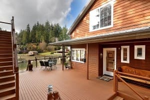 A wooden deck attached to a two-story house features patio furniture, a wooden bench, and overlooks a tree-lined lake. Nearby, stairs lead up to the second floor.