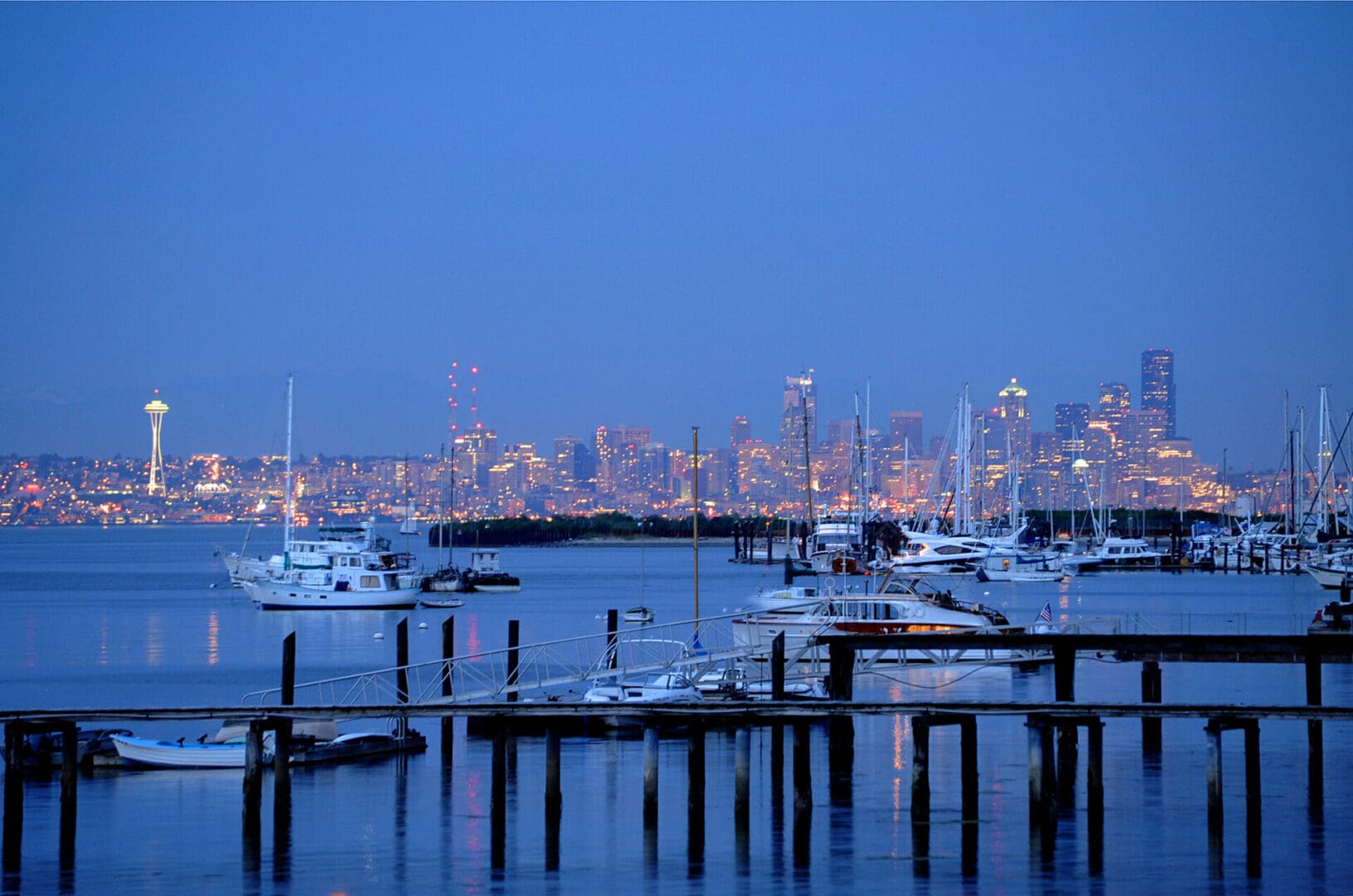 A marina with docked boats is in the foreground, and a city skyline, including a prominent tower, is visible in the background at dusk.