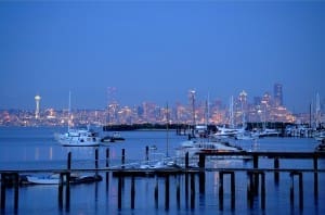 A marina with boats docked in the foreground, and a city skyline illuminated at dusk in the background.