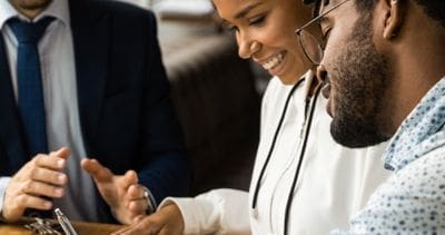 Three people sit at a table, two of them reviewing documents and writing with pens, while the third person gestures with their hands.