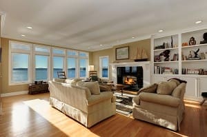 Spacious living room with large windows offering an ocean view, furnished with beige sofas, a fireplace, built-in shelves, and wooden flooring.