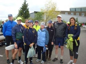 A group of nine people dressed in athletic gear stand outdoors, smiling at the camera. Some are wearing caps and jackets, with a building and tree in the background.