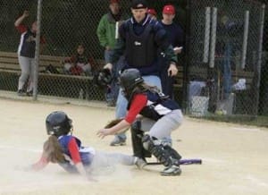 A softball player slides into home plate as the catcher attempts to tag them out while an umpire observes the play.