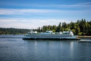 A large white and green ferry travels across a calm body of water with a forested shoreline and a clear blue sky in the background.