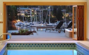 View from an indoor swimming pool overlooking a deck, with lounge chairs and a marina with boats docked on calm water, framed by sliding doors.