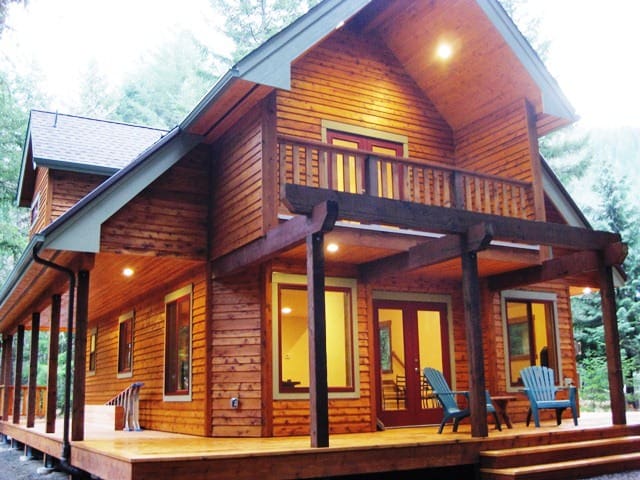 A two-story wooden cabin with a wraparound porch, pitched roof, and lit exterior lights sits amidst a forested background. Blue Adirondack chairs are placed on the porch.