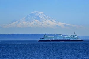 A ferry crosses a large body of water with a snow-capped mountain in the background under a clear blue sky.