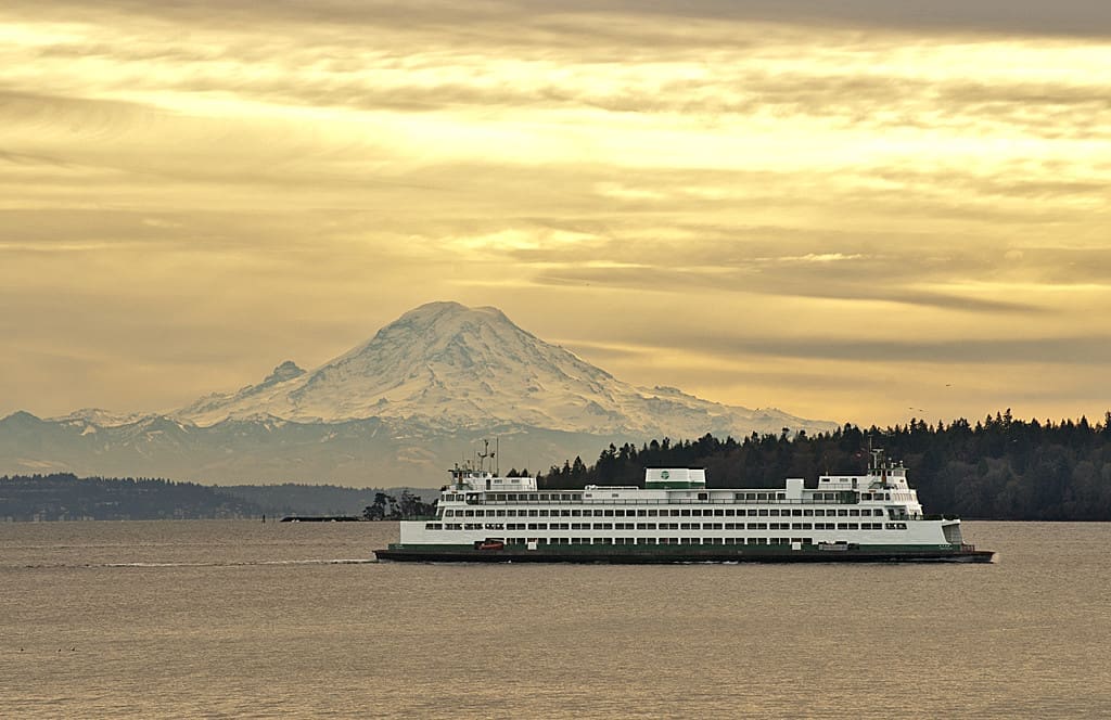 A ferry sails on calm waters against the backdrop of a towering snow-covered mountain under a cloudy sky.