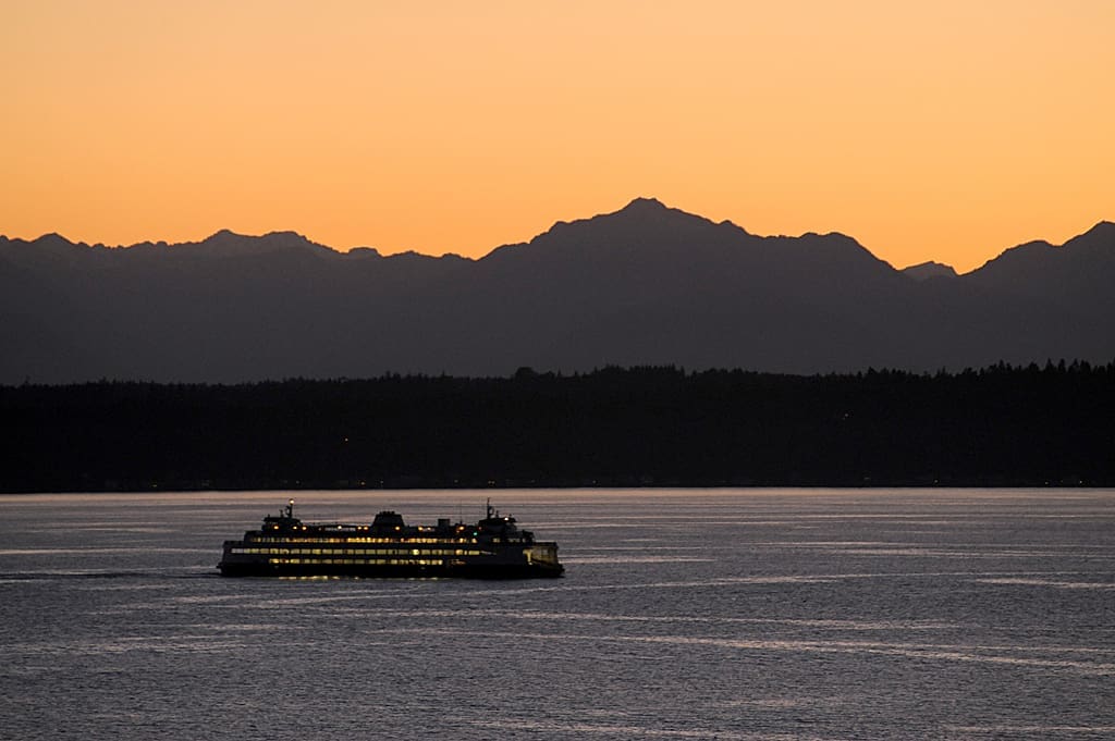 A ferry boat sails across a calm body of water at dusk, with silhouetted mountains in the background under an orange sky.