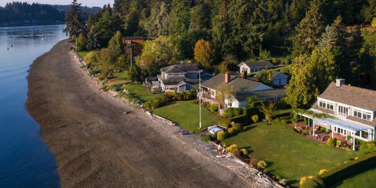 Aerial view of several houses with landscaped yards next to a forested area and a pebble beach along a calm body of water, perfect for showcasing in a market report.