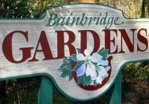 Sign reading "Bainbridge Gardens" with floral illustration, displayed outdoors against a backdrop of greenery.