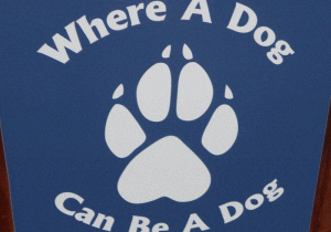 A sign with a white dog paw print on a blue background and the words "Where A Dog Can Be A Dog" displayed around it.