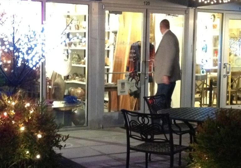 A man in a blazer stands at the entrance of The Island Gallery, a storefront decorated with lights and featuring a patio table and chairs and greenery in the foreground.