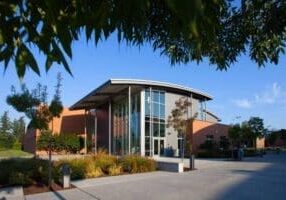 A modern building with large glass windows and a curved roof is surrounded by landscaped greenery on a sunny day.