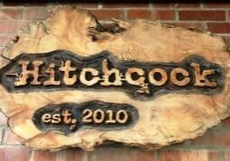 Wooden sign on a brick wall with the text "Hitchcock est. 2010" carved into it.