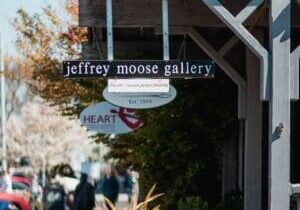 A street sign for "Jeffrey Moose Gallery" with an additional sign below indicating "Heart" and "Est. 1996." The area around is bustling with people and some trees are visible in the background.