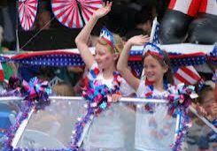 Two children wave from a decorated float during a parade. The float features patriotic colors and decorations, and the children are wearing festive headbands and leis.