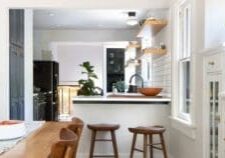 A modern kitchen with white walls, wooden shelves, and a black countertop. Two wooden bar stools are placed at the counter, and a wooden dining table with a bowl of food on it is in the foreground.