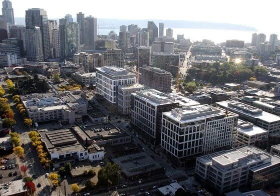 Aerial view of a cityscape with modern high-rise buildings, streets lined with trees showing fall colors, and a waterfront in the background under a bright sky.