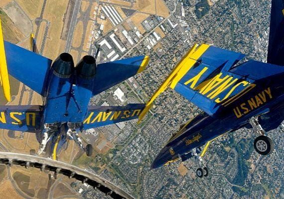 Two U.S. Navy Blue Angels jet aircraft fly upside down in close formation above a city landscape near an airfield.