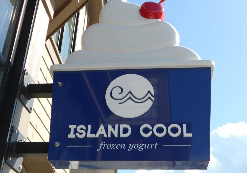 Blue and white "Island Cool frozen yogurt" sign with a swirl of frozen yogurt and a cherry on top, mounted on a building wall.