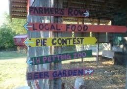 Colorful wooden signs on a farm point to various attractions: Farmers Row, Local Food, Pie Contest, 1H Petting Zoo, and Beer Garden.