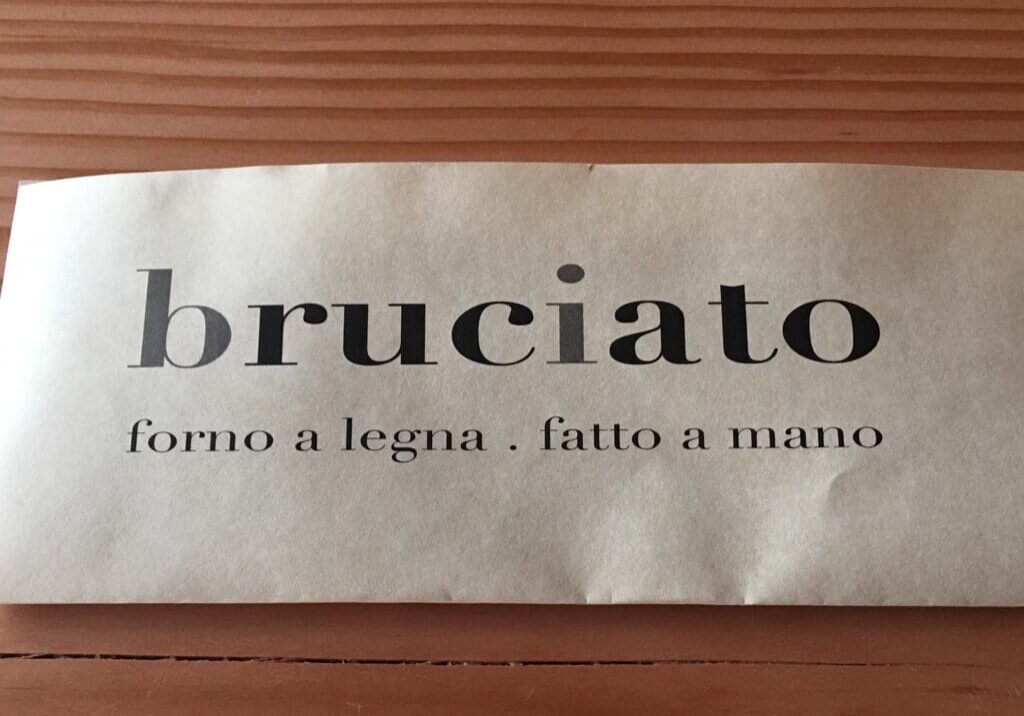 A paper with the text "bruciato forno a legna . fatto a mano" placed on a wooden surface.