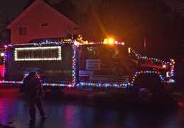 A fire truck decorated with colorful Christmas lights is parked on a wet street at night, with a dimly lit house in the background.