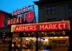 Neon sign reading "Public Market" and "Farmers Market" at an entrance with a clock and buildings in the background.