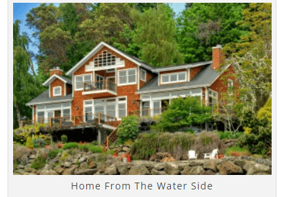 A large house with multiple stories, wooden exterior, and expansive windows. It is surrounded by trees and situated near a rocky waterfront. Two chairs and a small table are visible near the water.