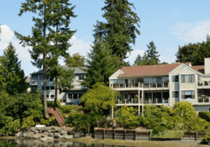 A scenic view shows a modern lakeside house with large windows and multiple balconies, surrounded by trees and greenery, reflecting on calm water under a clear blue sky.