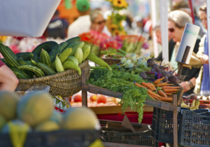 A bustling farmers' market with people browsing fresh vegetables including carrots, cucumbers, and radishes displayed in baskets and crates.