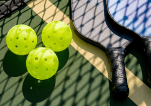 Two pickleball paddles and three yellow pickleballs are placed on a pickleball court, partially shadowed by the net.