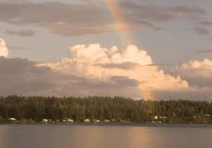 A rainbow arches over a calm lake with a forested shoreline in the background under a partly cloudy sky.