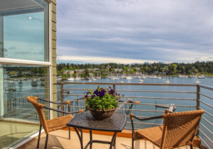 Balcony overlooking a lake with two wicker chairs, a table, and a potted plant. The water is calm with boats in the distance, and trees line the opposite shore.