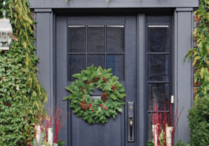 A dark blue front door decorated with a green wreath featuring pinecones, flanked by two planters with red twigs, white birch branches, and greenery. The doorway is framed with hanging greenery.