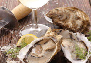 A wooden table with two glasses of white wine, a few opened oysters on the half shell, a lemon wedge, and a scattering of salt. An oyster knife is also visible.