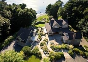 Aerial view of two houses with dark roofs surrounded by trees, near a body of water. A driveway and landscaped garden are visible between the houses.