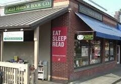 Street view of a brick building with two shops. The left shop is "Eagle Harbor Book Co." with a "Books" sign. The right shop has a large red sign that reads, "EAT SLEEP READ." Blue awning above windows.