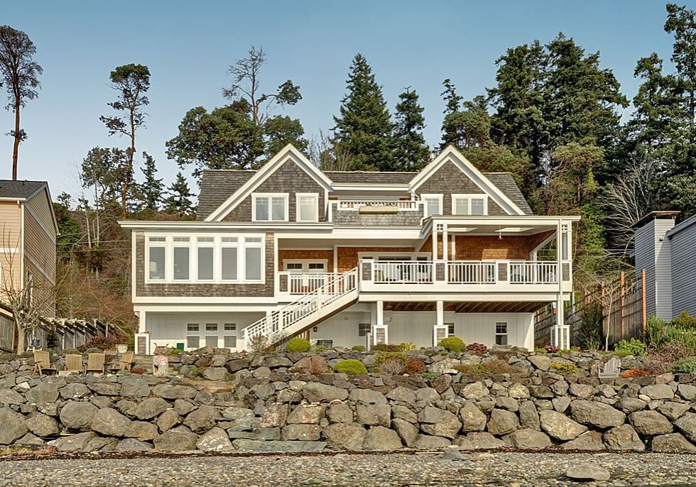 A two-story house with large windows and a spacious deck, surrounded by trees and built on a stone retaining wall.