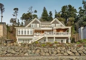 Two-story house with large windows, white railings, and a stone retaining wall in front, surrounded by trees.