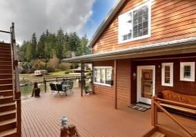 A wooden deck attached to a two-story house features patio furniture, a wooden bench, and overlooks a tree-lined lake. Nearby, stairs lead up to the second floor.