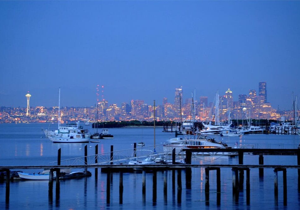 A marina with docked boats is in the foreground, and a city skyline, including a prominent tower, is visible in the background at dusk.