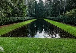 A rectangular reflecting pool is surrounded by neatly trimmed grass and bordered by hedges and tall trees.