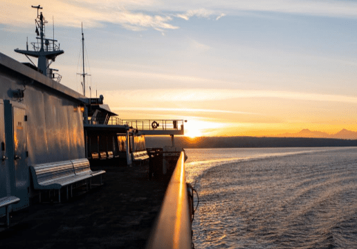 A metal deck of a ferry is shown at sunset, with empty benches and railing leading the eye toward the horizon. The sky features a blend of blue and orange hues above a body of water.