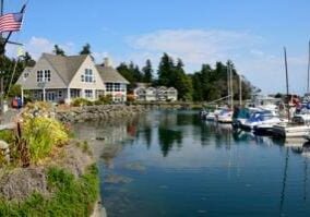 A waterside house with an American flag and docked boats on a calm body of water. Other houses and trees are in the background under a clear blue sky.