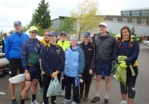 A group of nine people dressed in athletic gear stand outdoors, smiling at the camera. Some are wearing caps and jackets, with a building and tree in the background.