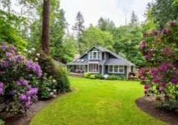 A quaint house with a pitched roof and dormer windows is surrounded by a lush green lawn and colorful flowering bushes, set in a wooded area with tall trees.